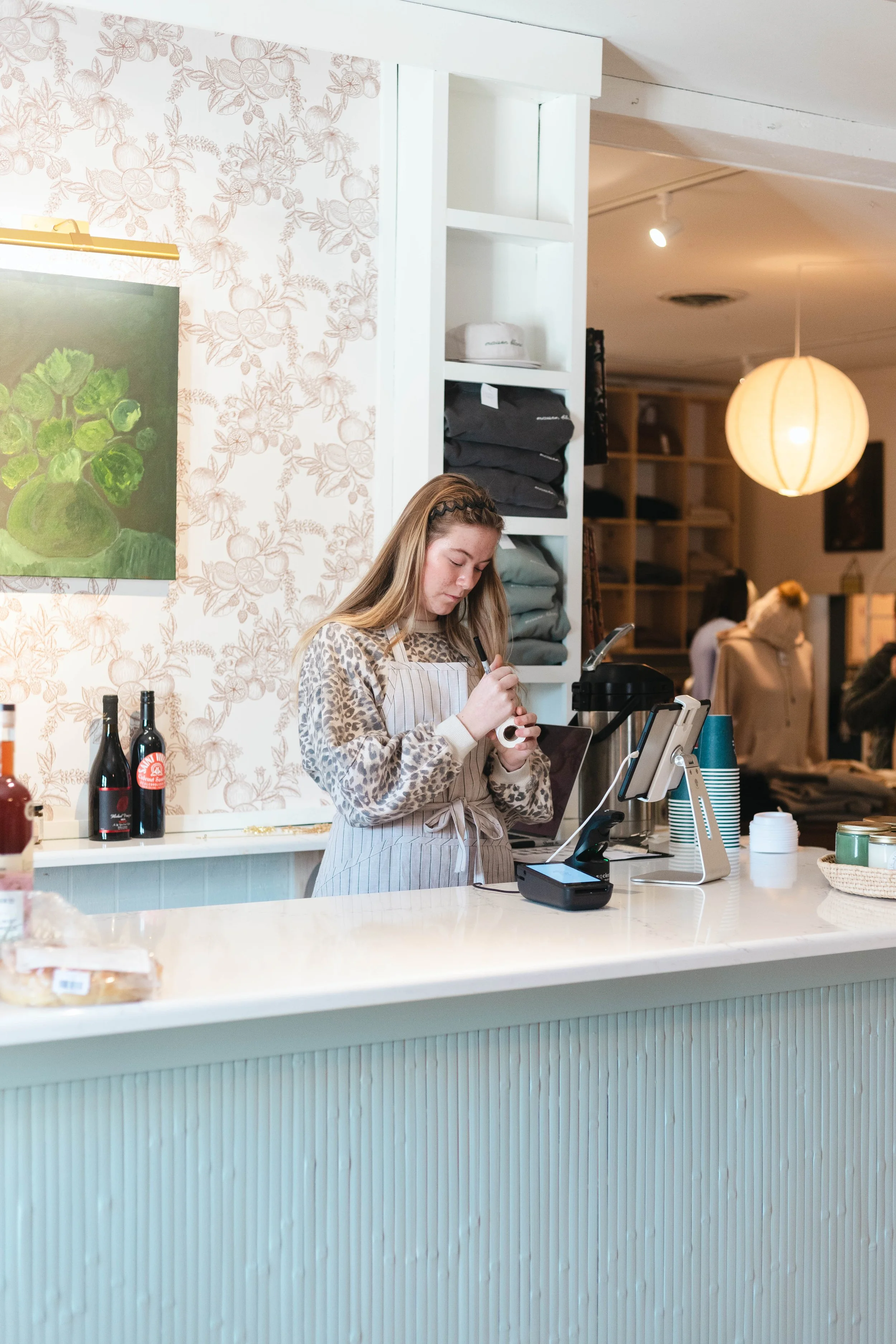 A young female cashier with a blonde ponytail and a leopard print sweater, wearing a beige apron, working at a cafe counter using a payment terminal.