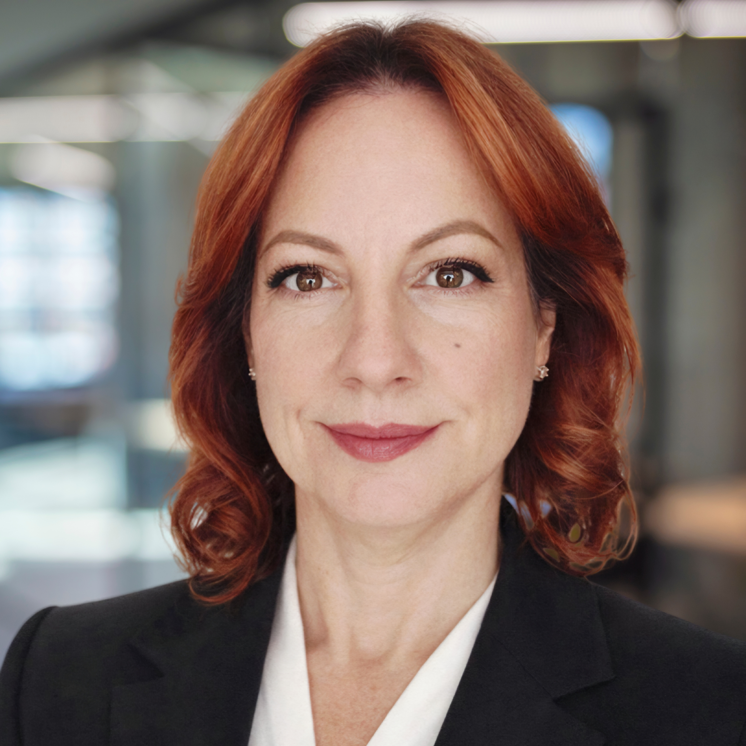 A professional woman with medium-length red hair, wearing a black blazer and white blouse, smiling against a warm beige background.