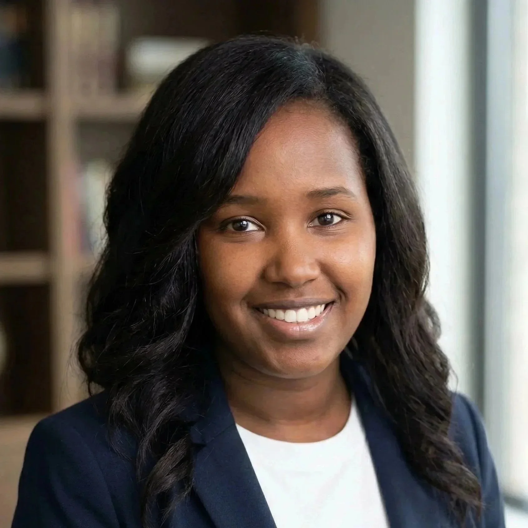 Close-up of a woman with black, wavy hair, smiling, wearing a white shirt and a navy blazer, standing indoors near a window.