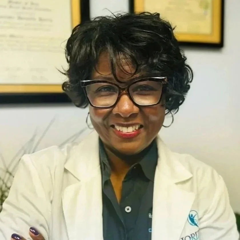 Portrait of a confident African American female healthcare professional wearing glasses, smiling, dressed in a white coat with a badge, standing against a blurred background of framed documents.