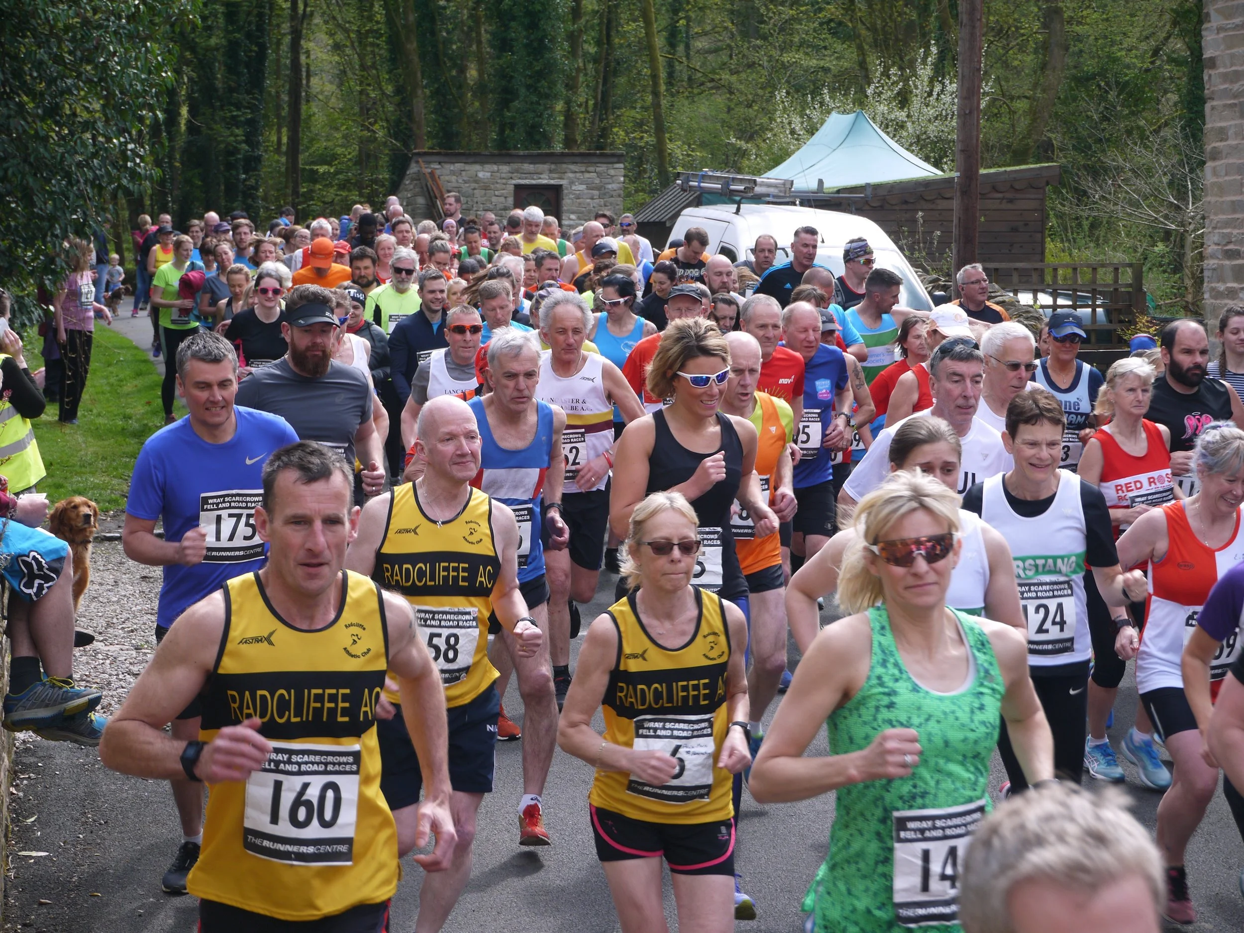 Wray Scarecrow Festival; Crowd of runners participating in a race outdoor on a paved path surrounded by greenery and trees, with a white van and structures in the background