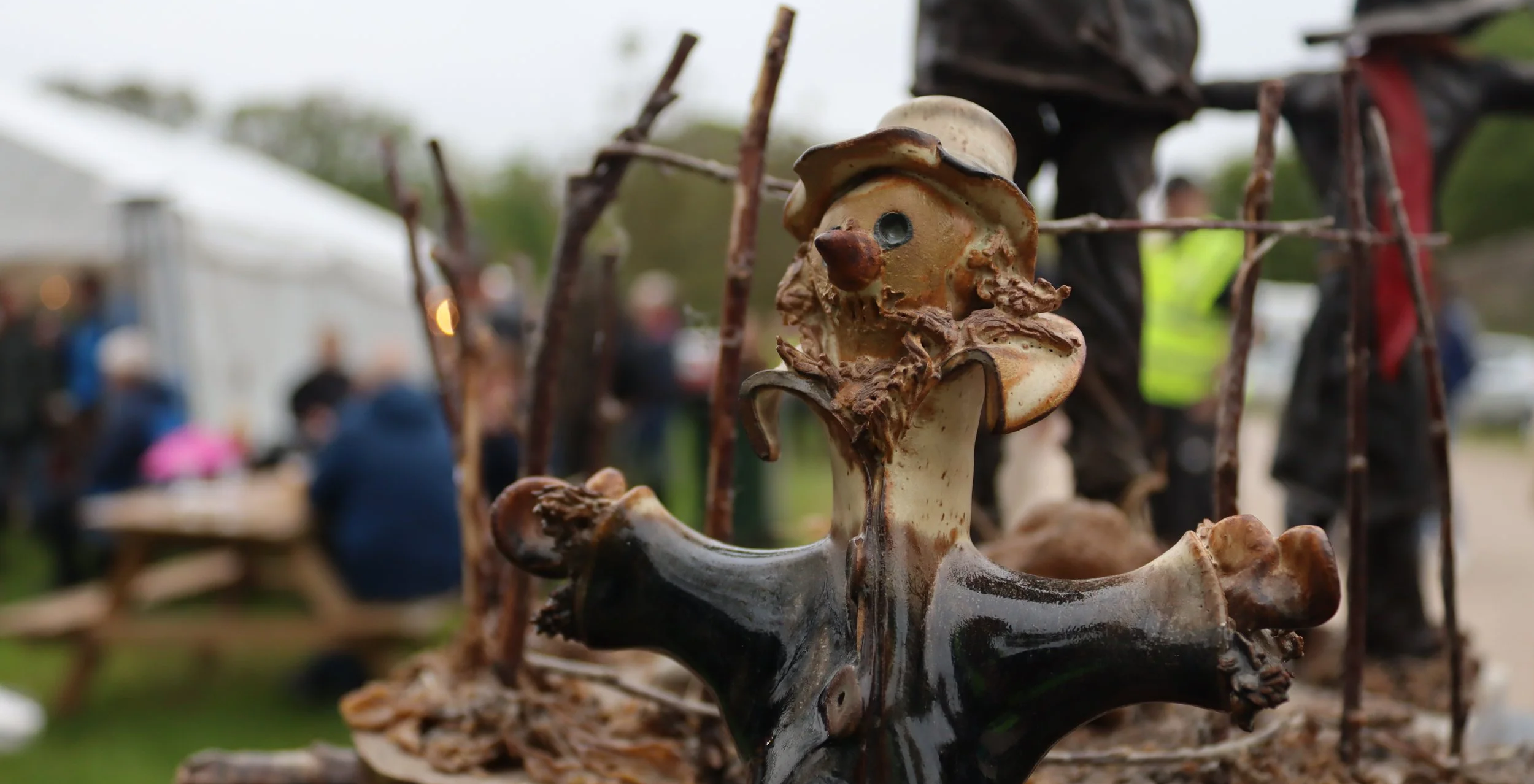 A close-up of a mushroom sculpture with a face, wearing a hat, surrounded by sticks, at outdoor market with people in the background.