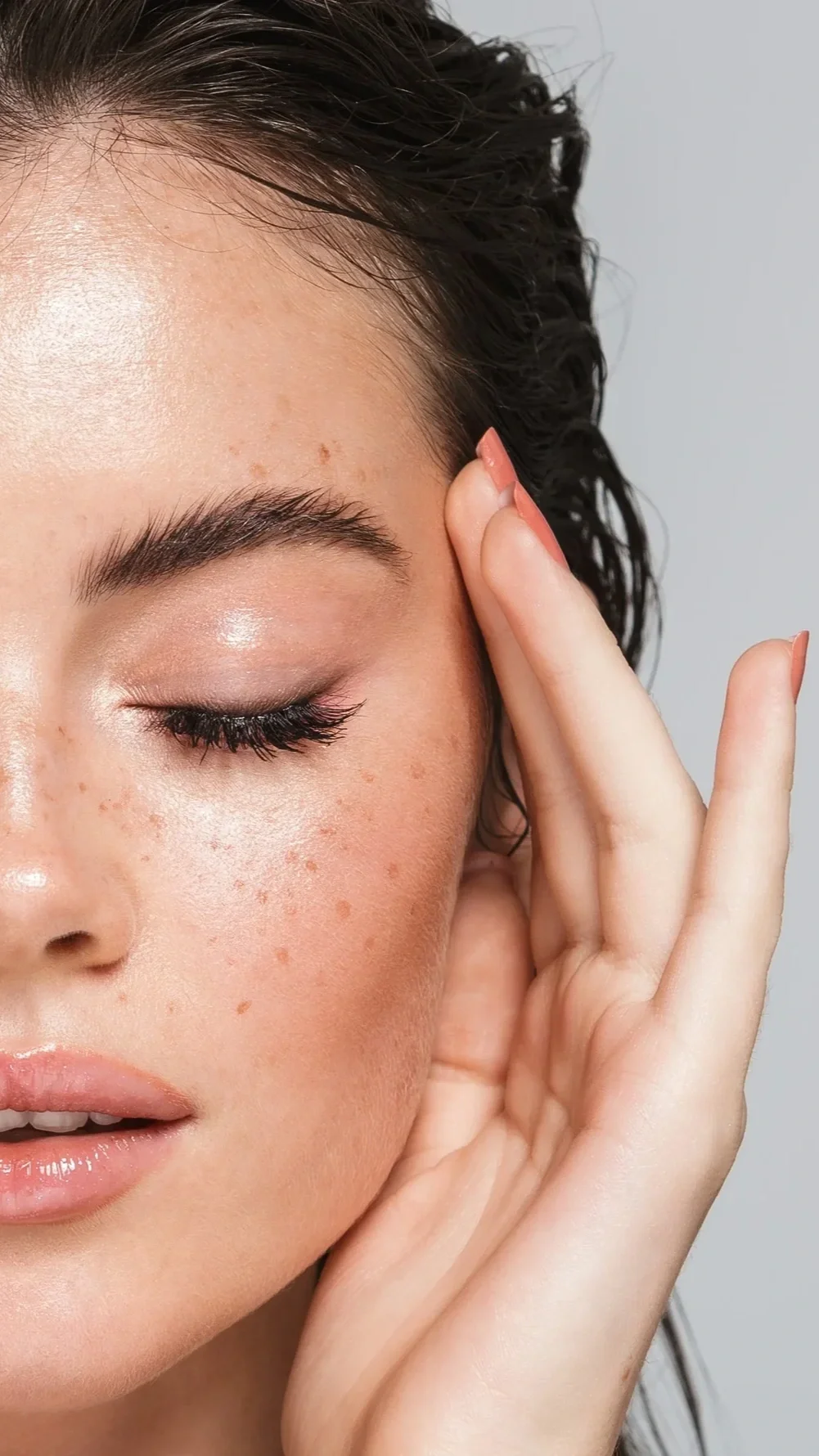 Close-up of a woman with closed eyes, freckles, and glossy lips, gently touching her temple with her fingers.