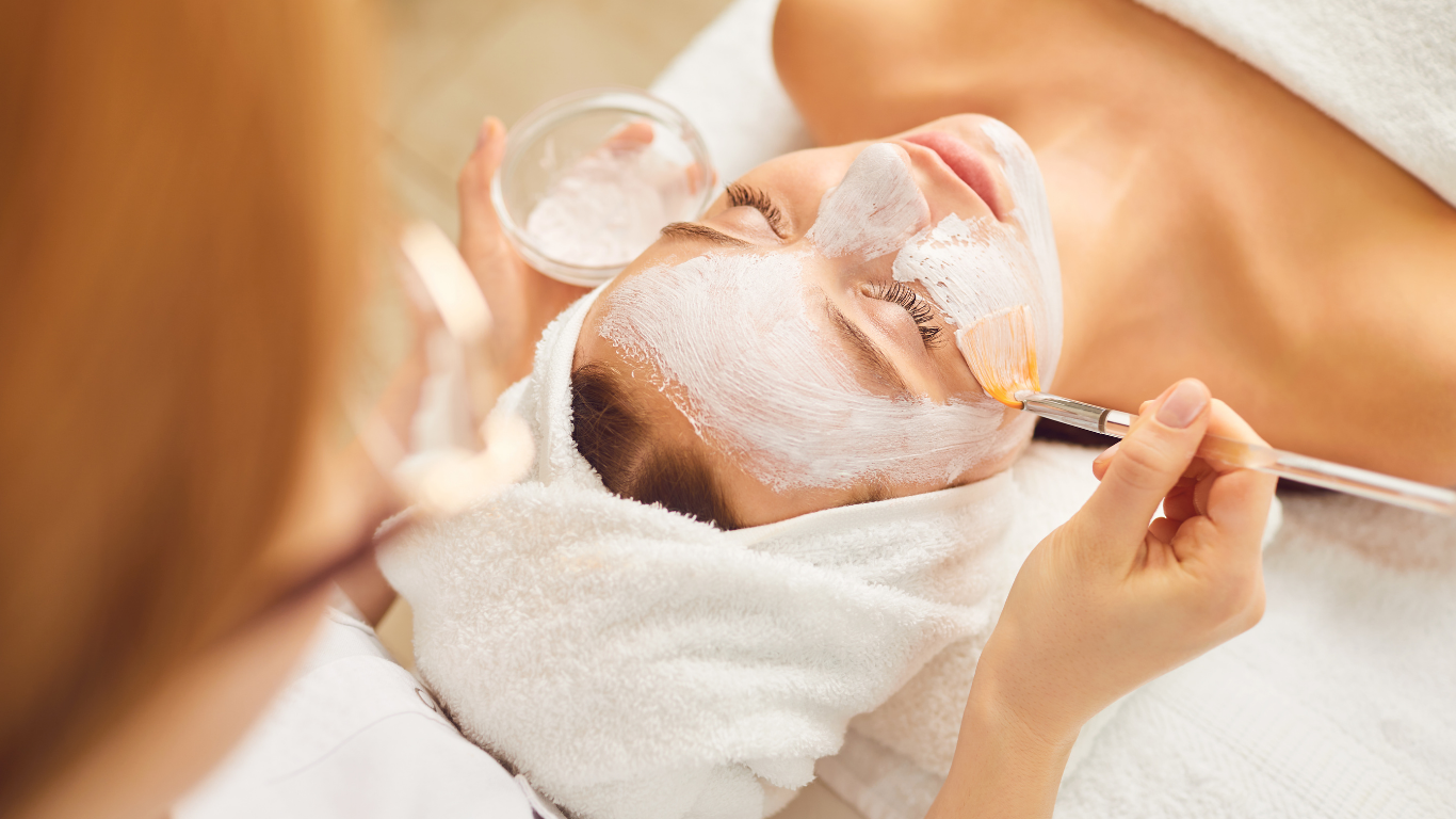 A woman lying down with her eyes closed, having a facial mask applied by an esthetician with a brush, in a spa setting.
