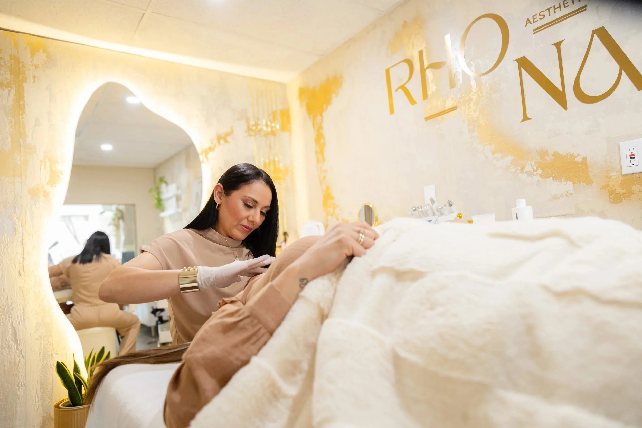 A woman with dark hair and earrings getting a tattoo on her arm at a tattoo studio with warm yellow lighting and a textured yellow wall with gold lettering that reads 'RHO AESTHETIC STUDIO'.