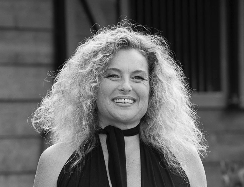 Black and white photo of a smiling woman with curly blonde hair, wearing a dark sleeveless top and a black scarf around her neck, standing outdoors in front of a wooden building.