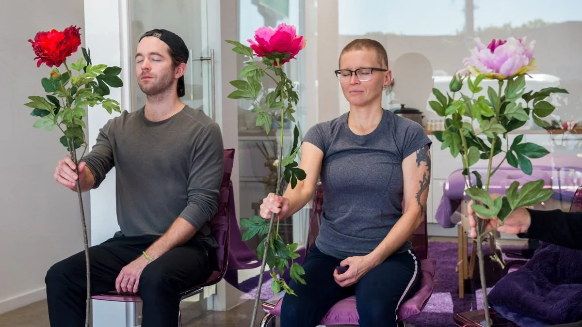 Three people sitting on purple chairs with closed eyes and meditative expressions, each holding large colorful flowers. Kismet is an intuitive living institute located in Venice, California, offering workshops, healing sessions, and spiritual educati