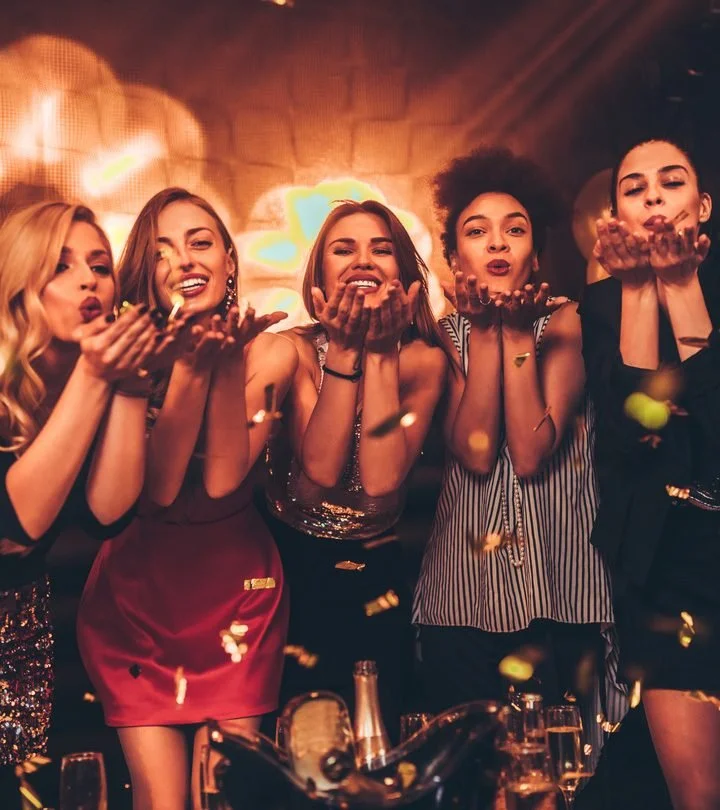 Five women cheer and blow kisses at a celebration or party, with confetti floating in the air and a warm, festive background. Kismet is an intuitive living institute located in Venice, California, offering workshops, healing sessions, and spiritual e