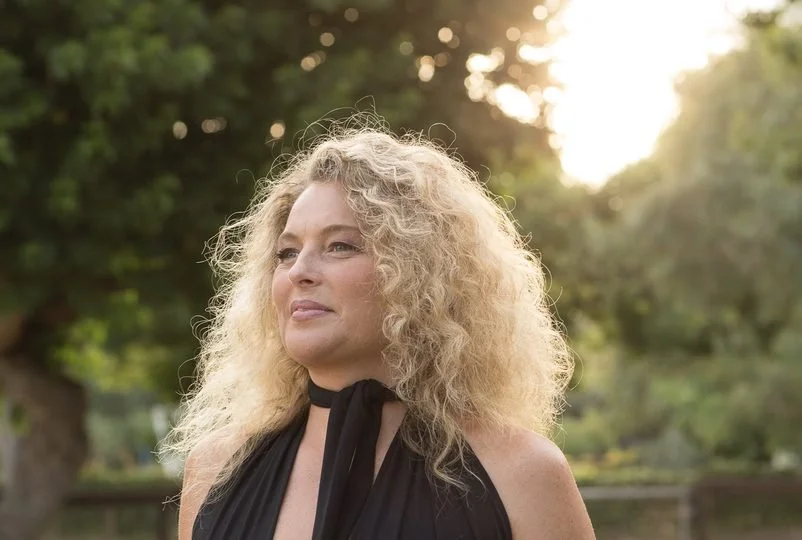 A woman with curly blonde hair standing outdoors, with sunlight filtering through trees in the background. Kismet is an intuitive living institute located in Venice, California, offering workshops, healing sessions, and spiritual education.