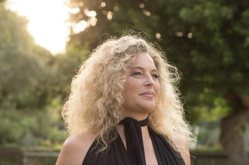 A woman with curly blonde hair standing outdoors in a park-like setting, wearing a black top and looking to the side. Kismet is an intuitive living institute located in Venice, California, offering workshops, healing sessions, and spiritual education