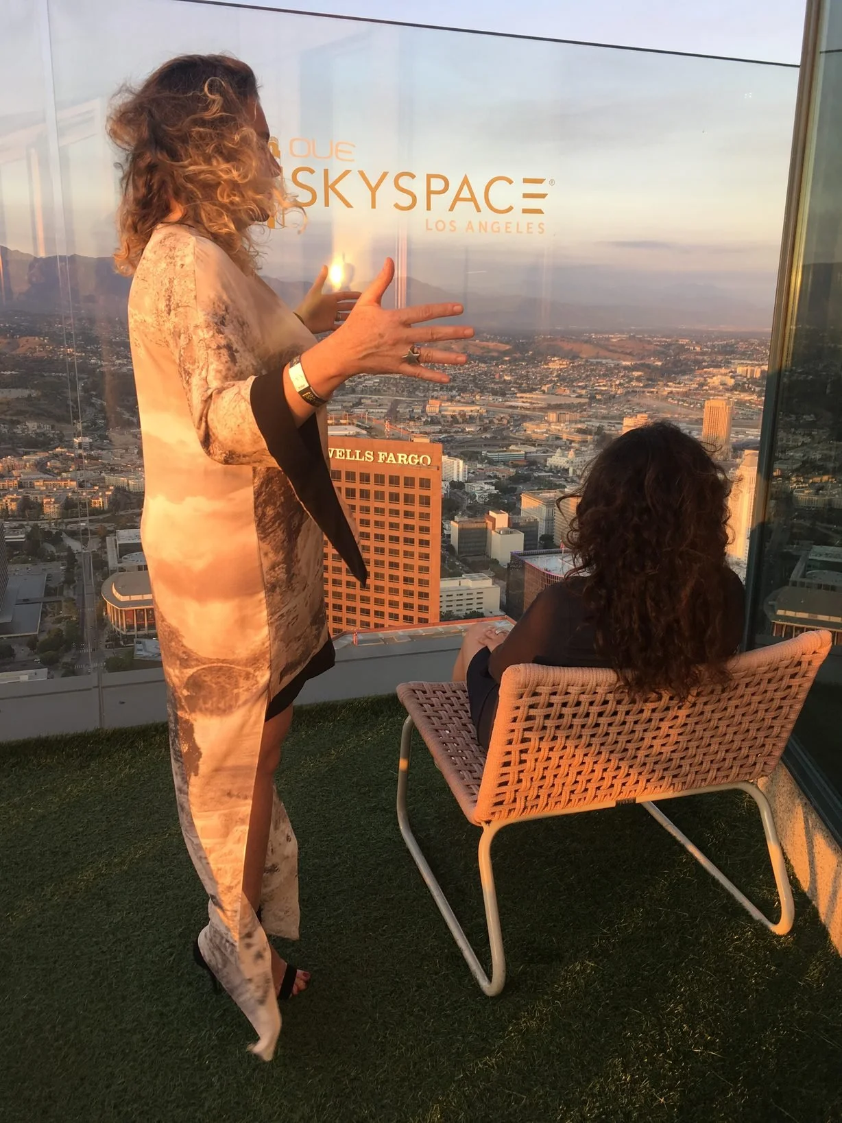 Two women on an outdoor balcony with a glass wall, overlooking the Los Angeles skyline at sunset. One woman is standing and gesturing with her hands, while the other is seated in a woven chair, looking out over the city. Kismet is an intuitive living
