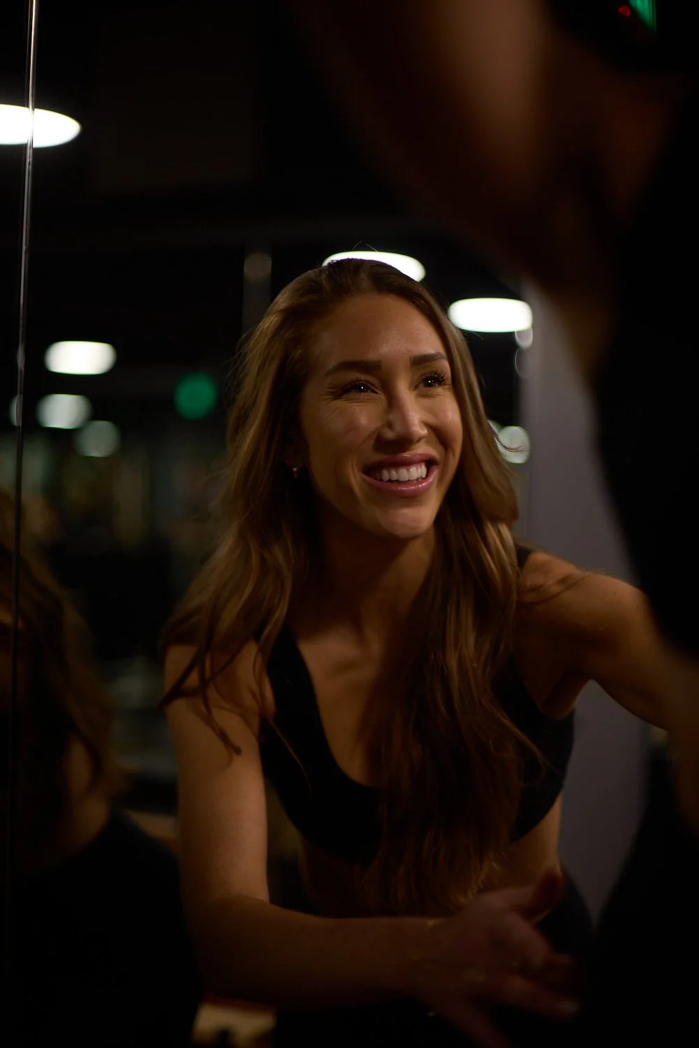 A smiling woman with long red hair is looking at her reflection in a mirror, wearing a black top in a dimly lit environment.