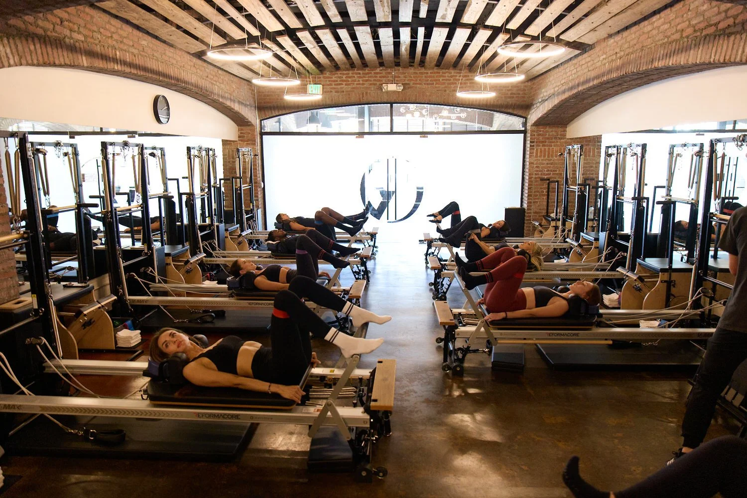 Women in a Pilates studio named Reformacore, exercising on reformer machines, with brick walls and large windows in the background.