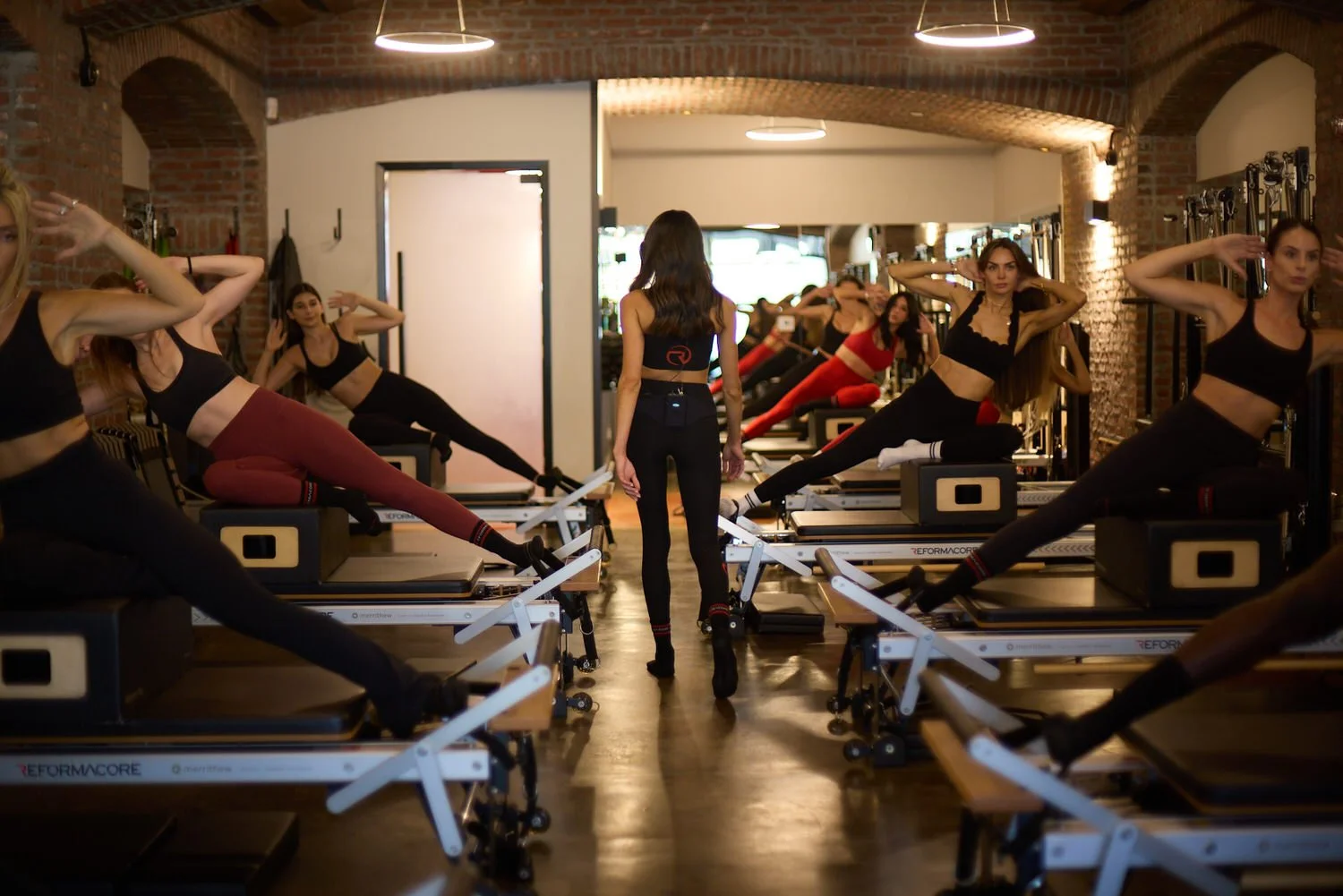 A group fitness class at Reformacore with women doing Pilates on reformer machines in a studio, led by an instructor walking in the center.