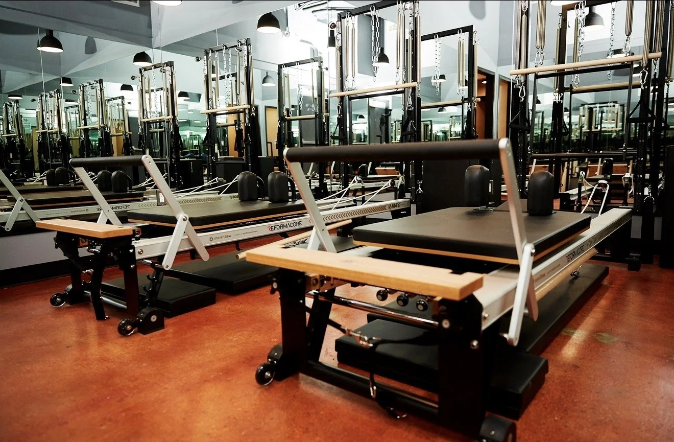A woman is exercising on a reformer Pilates machine at Reformercore in West LA. she is smiling while holding a circular Pilates ring. Fitness equipment surrounds her, and large windows provide natural light.