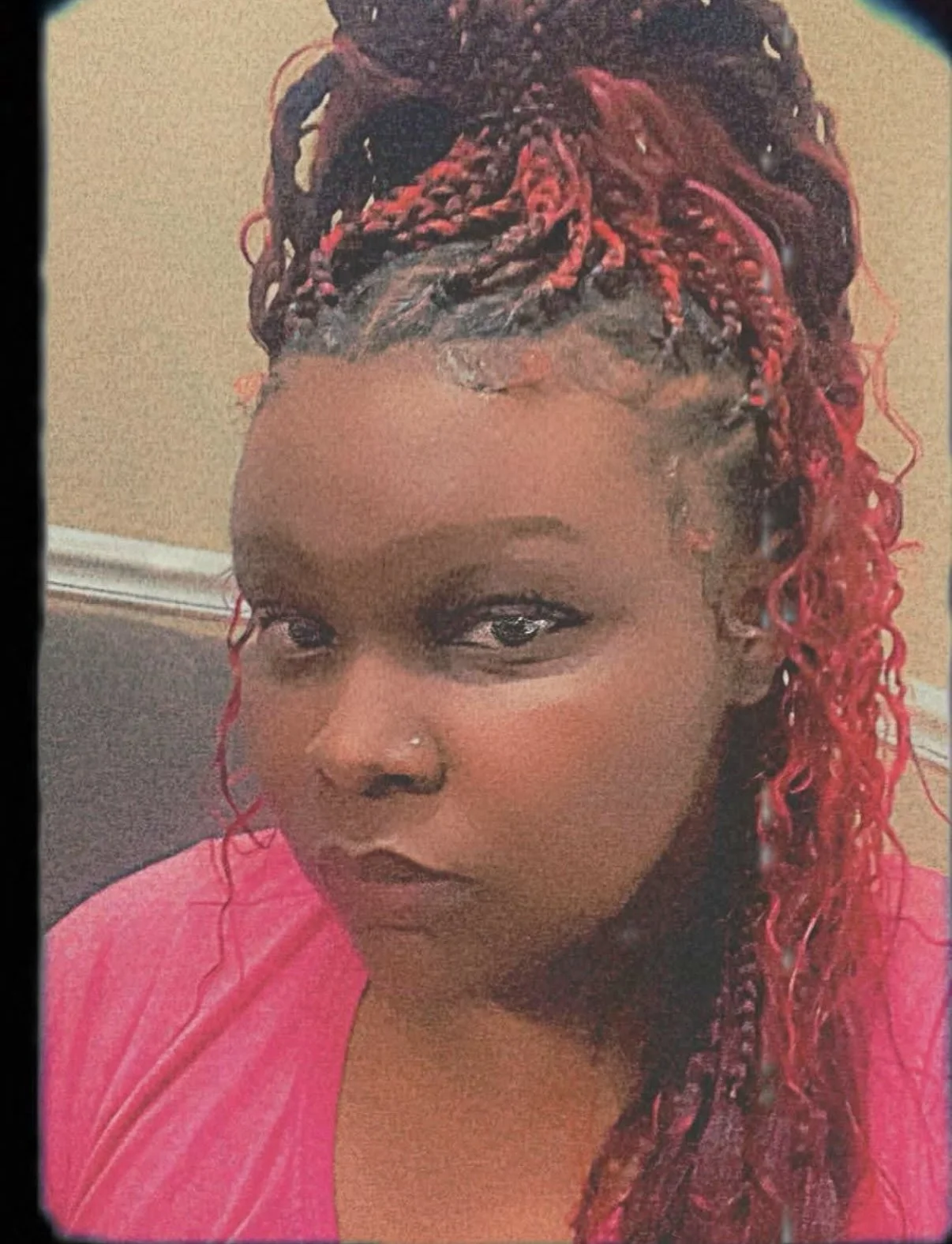 Close-up photo of a woman with dark skin, green eyes, and red and black braided hair cascading over her shoulder, wearing a pink top.