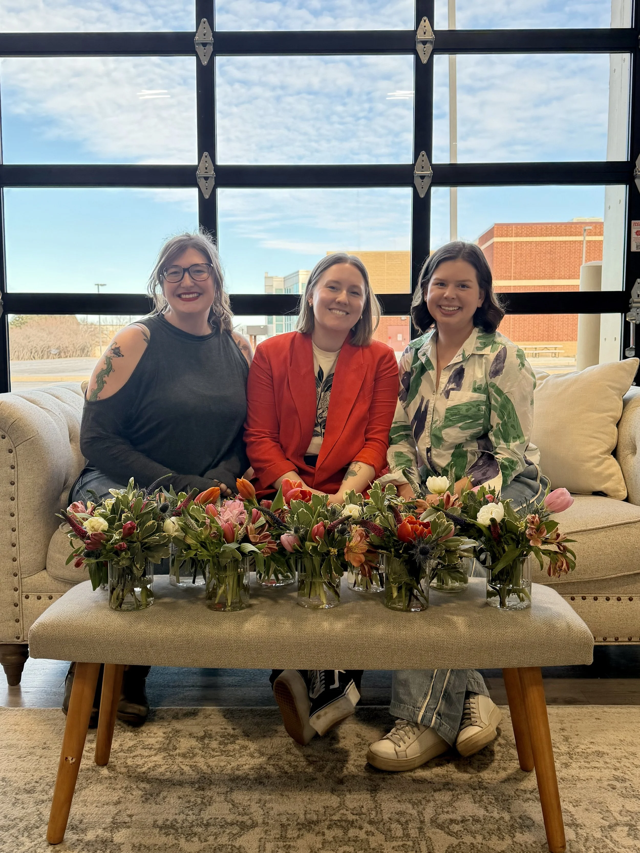 Three women sitting on a sofa behind a table with colorful flower arrangements in vases, large window with cloudy sky outside, and brick buildings in the background.