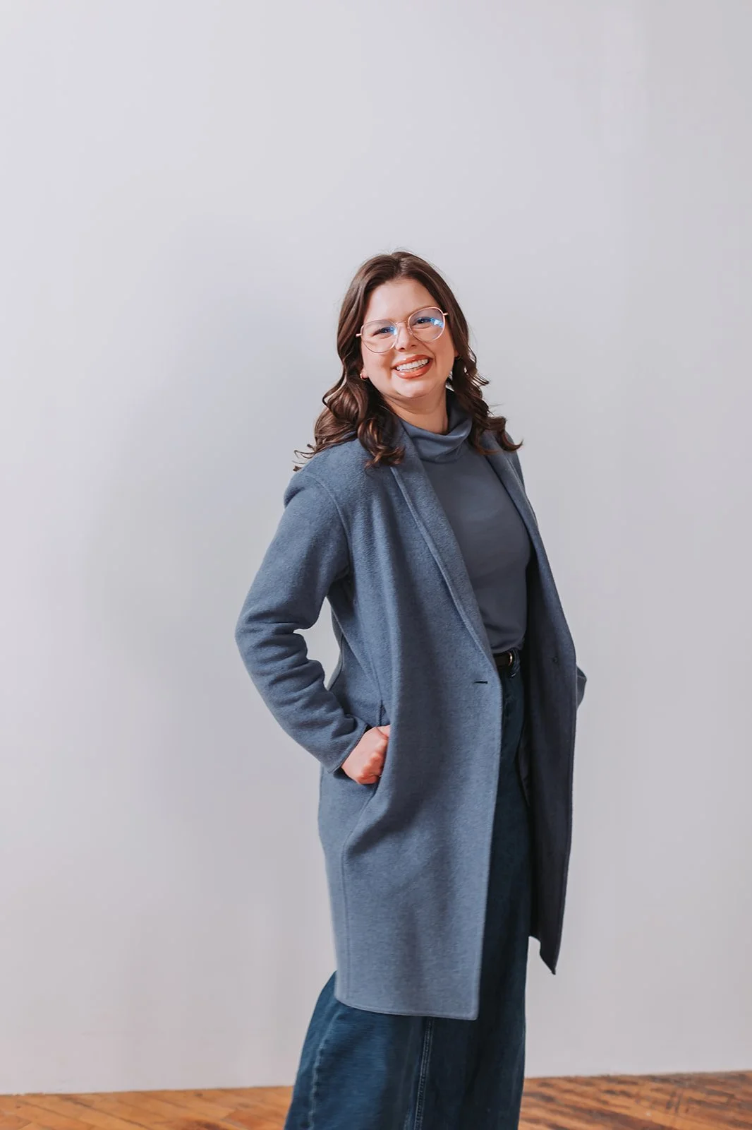 A woman standing indoors, smiling, wearing glasses, a blue turtleneck, a gray coat, and dark jeans, against a plain light-colored wall.