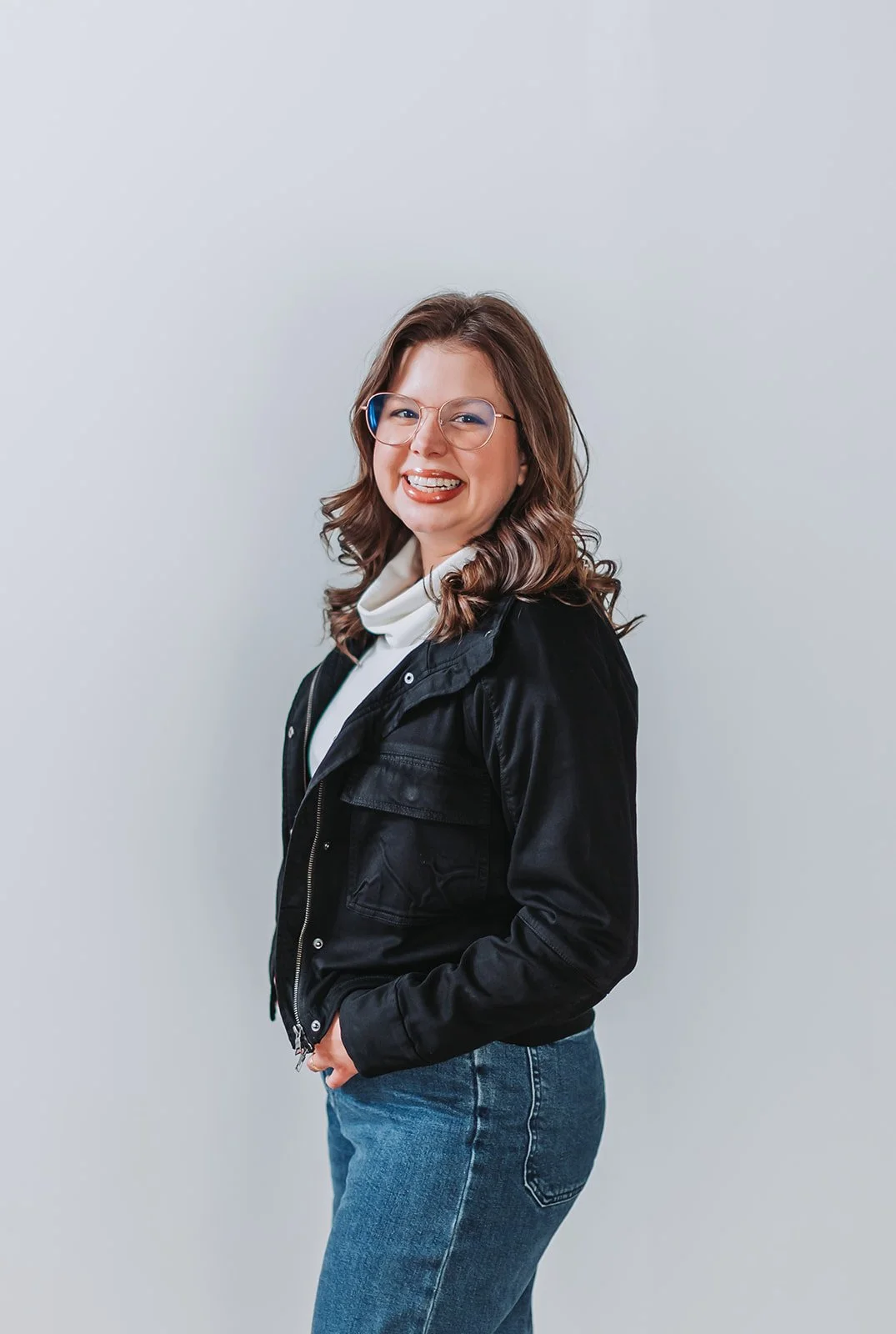 A young woman with wavy brown hair, glasses, and a big smile, wearing a black jacket, white shirt, and blue jeans, standing against a plain light-colored background.