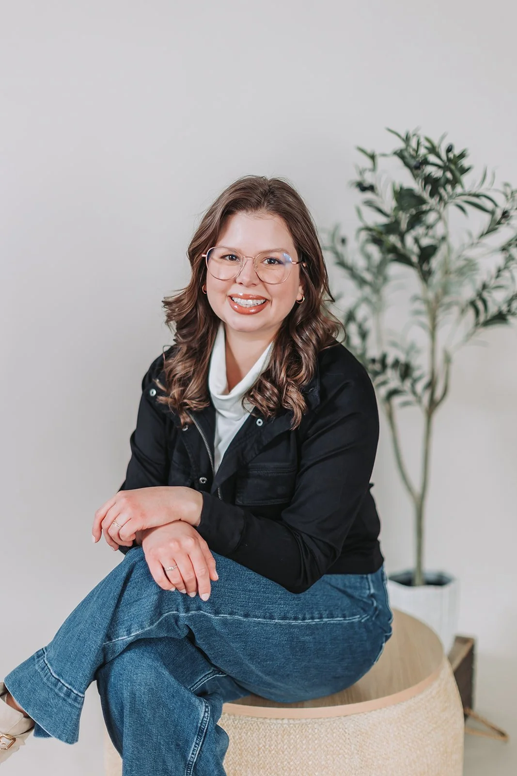 A woman with shoulder-length wavy brown hair, wearing glasses, a black jacket, a cream-colored turtleneck, and blue jeans, sitting on a round beige ottoman in front of a potted plant and white wall, smiling at the camera.