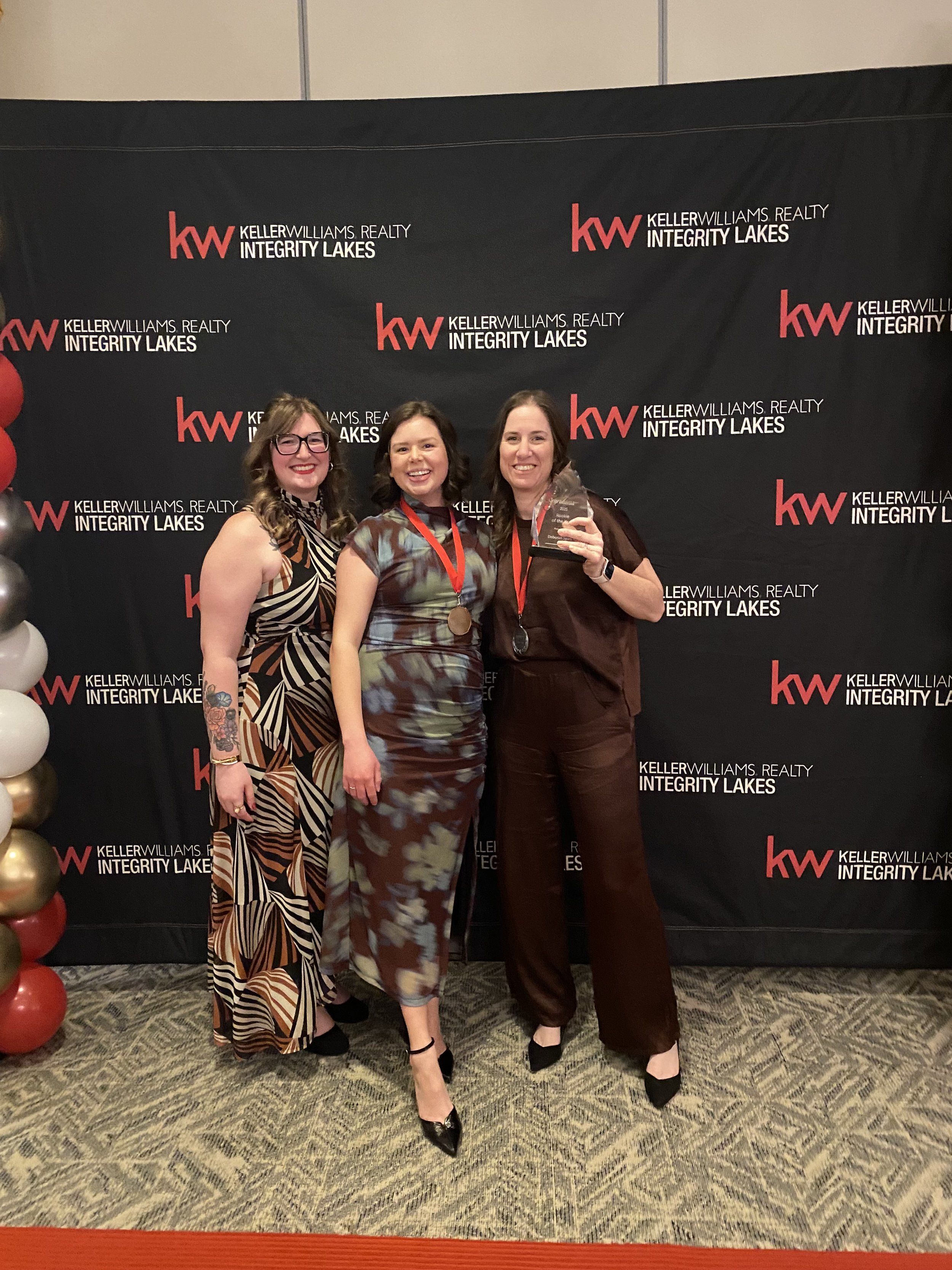 Three women standing together at a Keller Williams Realty event, smiling, with a black backdrop that reads Keller Williams Realty Integrity Lakes. The woman on the right is holding an award, and the women are dressed in stylish, patterned clothing.
