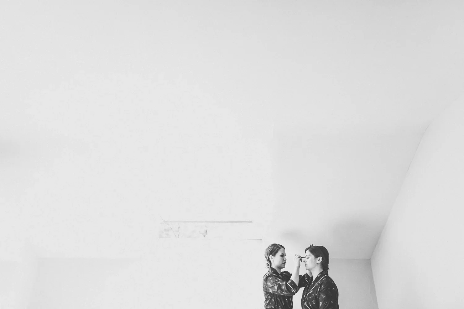 Two women in robes with floral patterns, one applying makeup to the other's face, in a minimalist room with a plain ceiling.