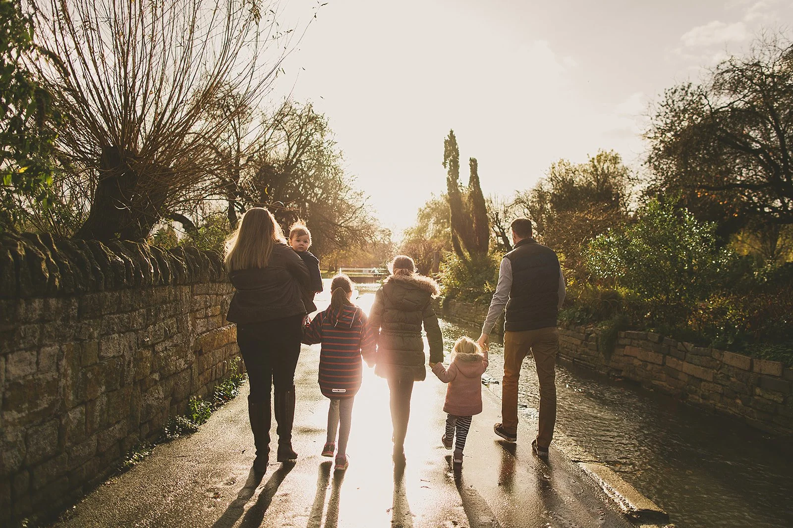 A family of seven walking along a paved path beside a stone wall, holding hands. The scene is bathed in warm sunlight, with trees on either side and a body of water visible in the background.