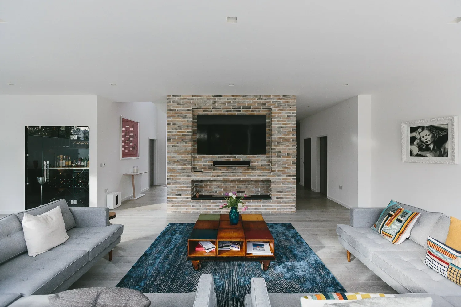 Living room with white walls, hardwood floors, a brick accent wall with a mounted flat-screen TV, and a coffee table with a blue vase of pink flowers. Two sofas with colorful pillows, and artwork on the walls.