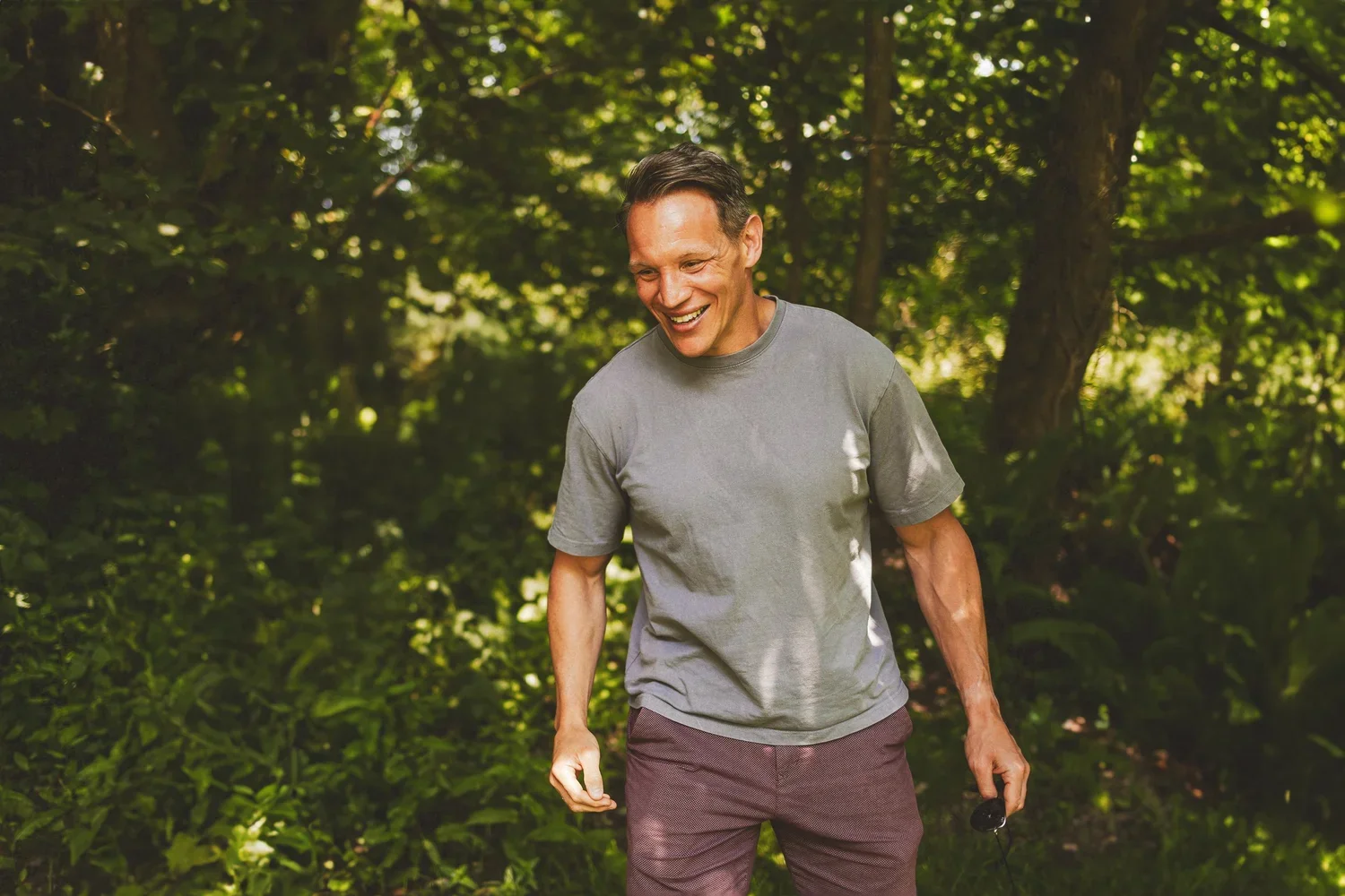 A man smiling and walking outdoors in a lush green forest, wearing a gray t-shirt and brown shorts, holding an object in his right hand.