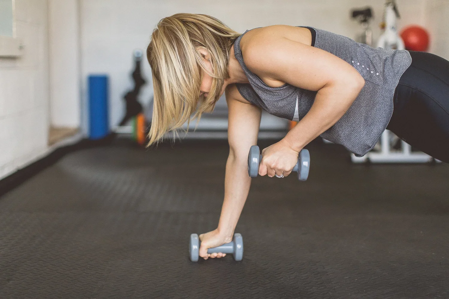 A woman with blonde hair exercising in a gym, doing a plank with dumbbells, supporting her body with one arm while holding a dumbbell in the other.