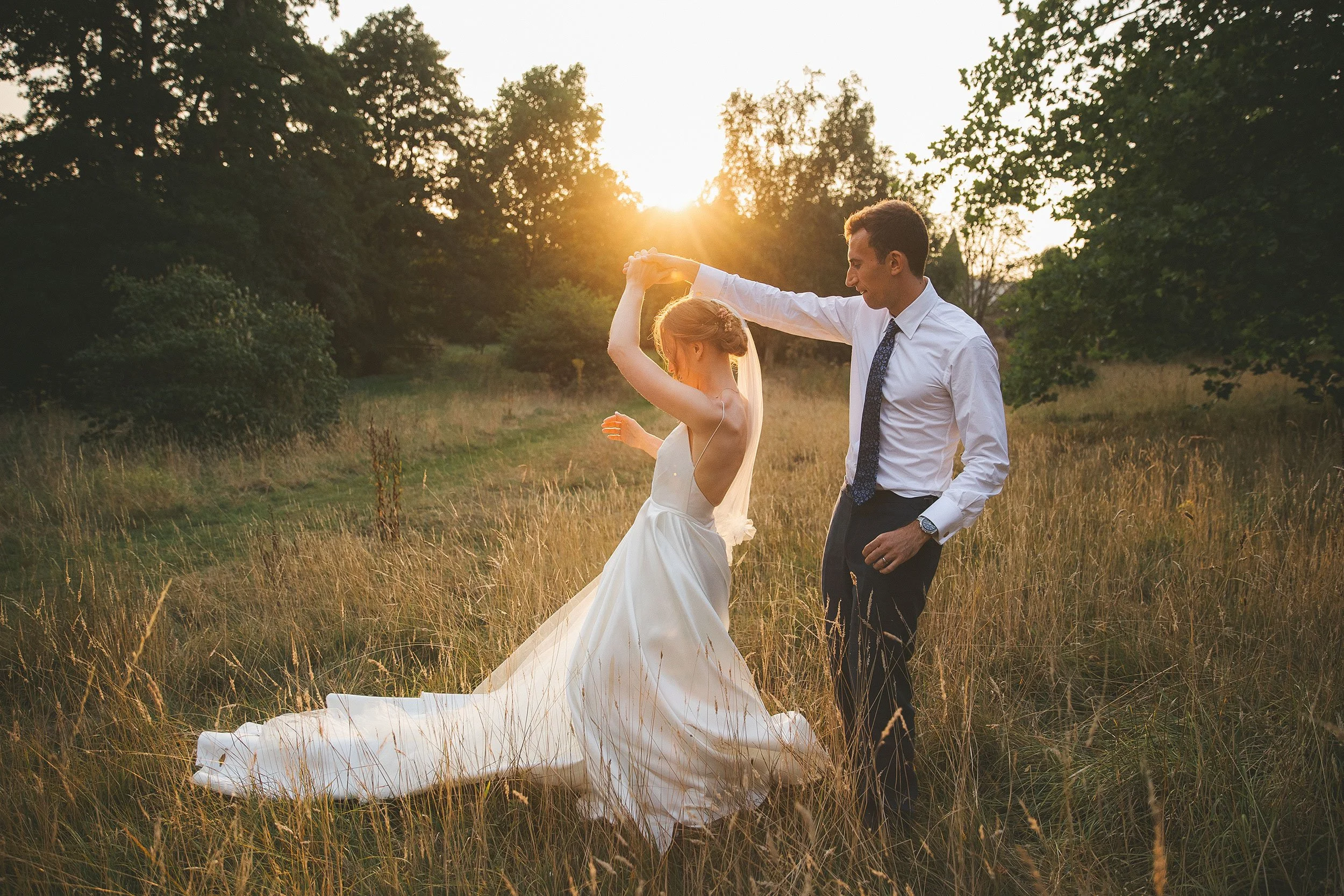 A bride and groom dancing outdoors in a grassy field during sunset, surrounded by trees.