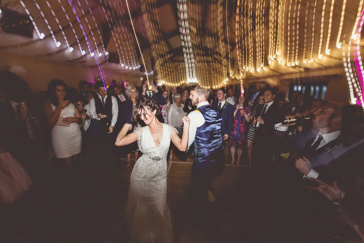 People dancing and celebrating at a wedding reception with colorful string lights overhead.