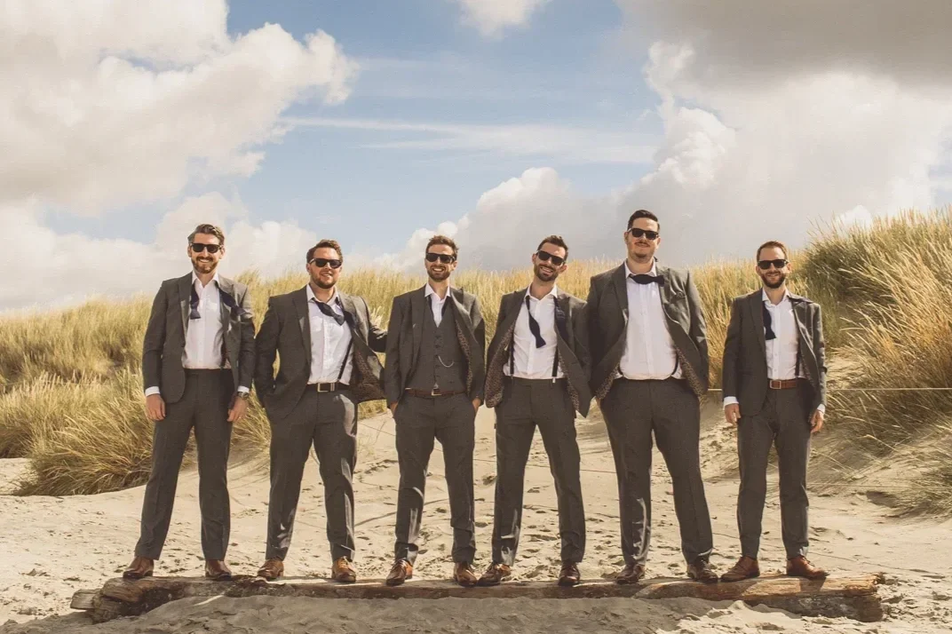 Six men in suits standing on a sandy beach with dunes and grass in the background, smiling and posing for a photo.