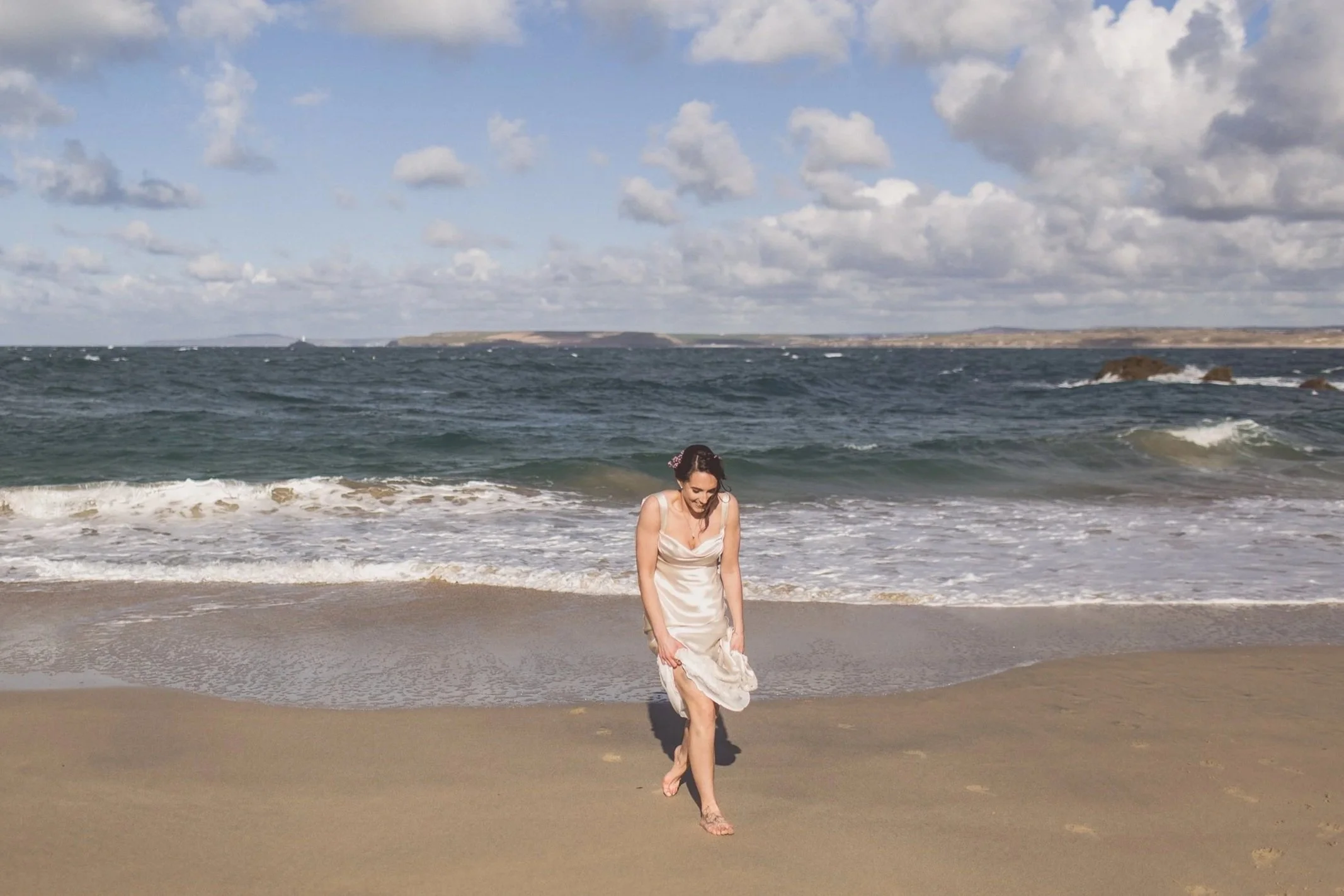 A woman in a white satin dress walks along the sandy beach near the shoreline with the ocean and a partly cloudy sky in the background.