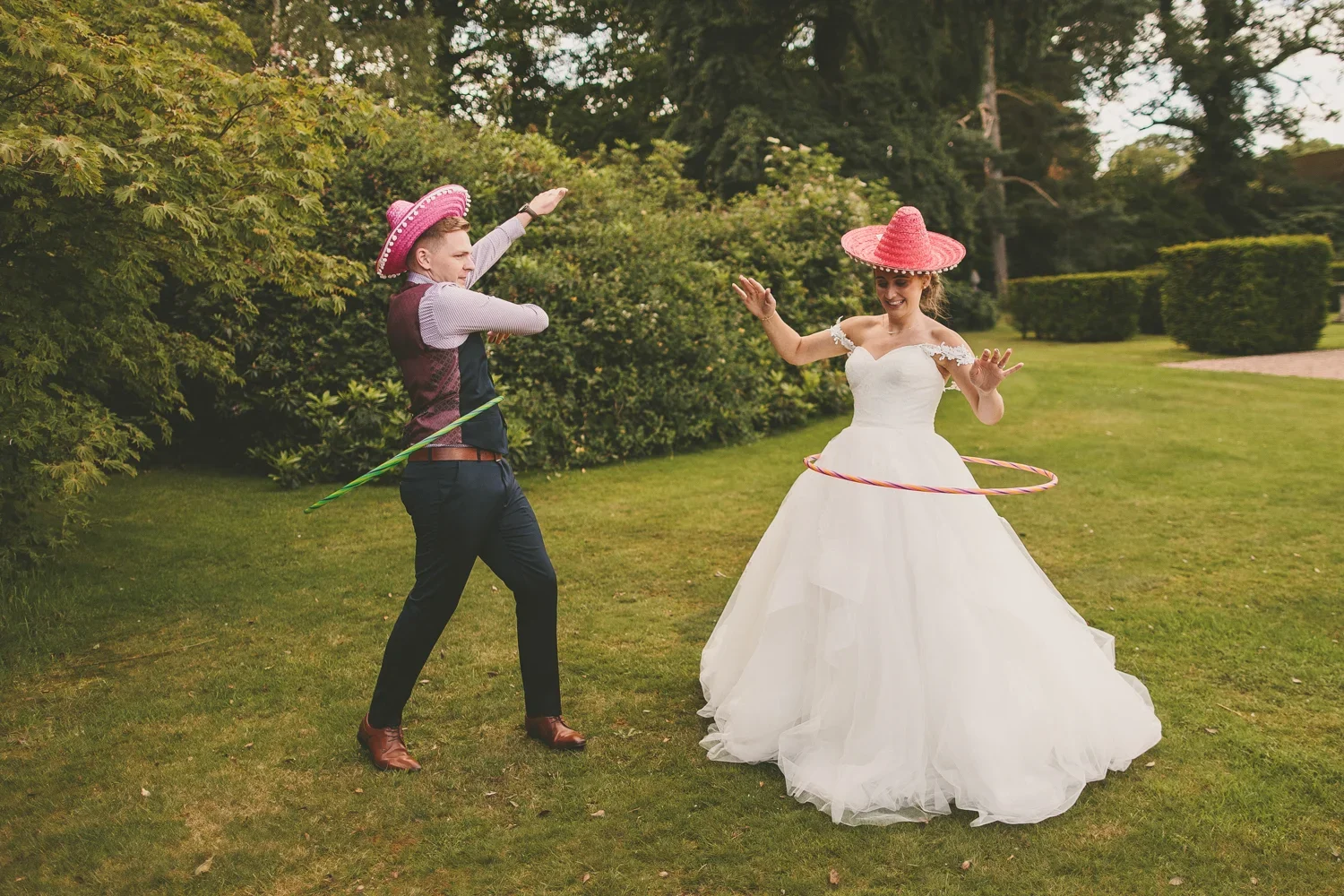A bride and groom in wedding attire playing with hula hoops outdoors on a grassy lawn, surrounded by trees and bushes on a cloudy day.