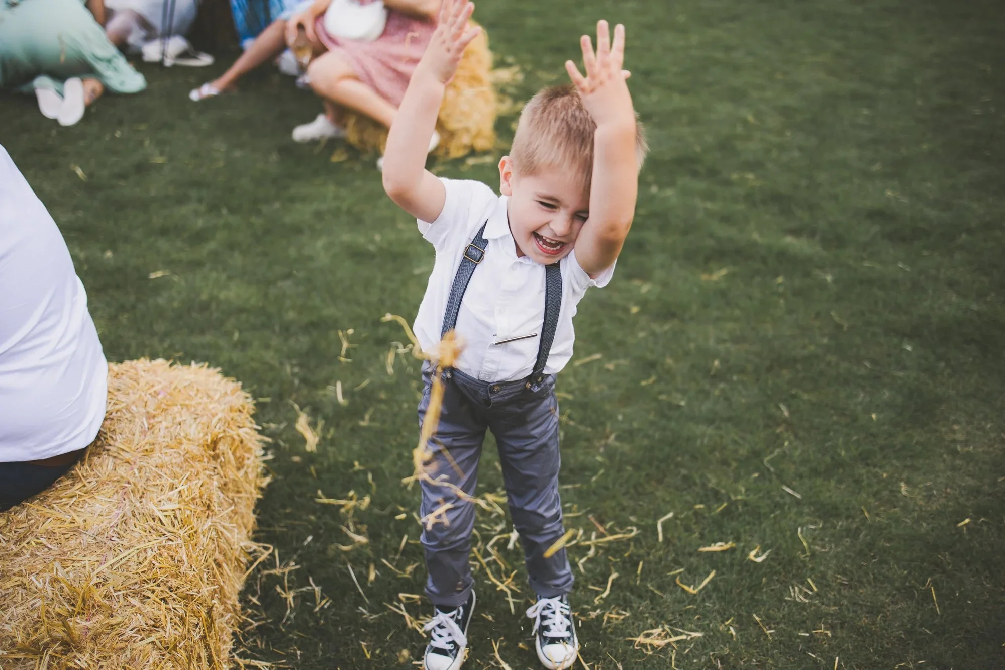 A young boy with blond hair, wearing a white shirt, black suspenders, and gray pants, is smiling and dancing in a grassy field. Behind him, a girl in a pink dress is sitting on the ground, and other children are partially visible nearby. A hay bale is in the foreground, and the children appear to be celebrating or playing outdoors.