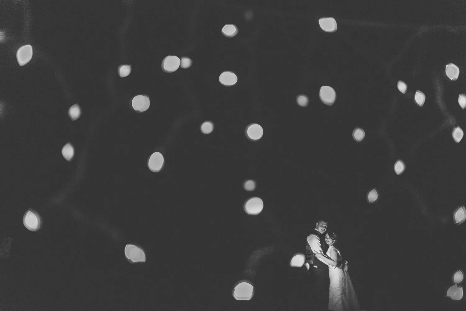 Black and white photo of a couple, possibly at a wedding, embracing and smiling at each other, with a backdrop of out-of-focus lights creating a bokeh effect.