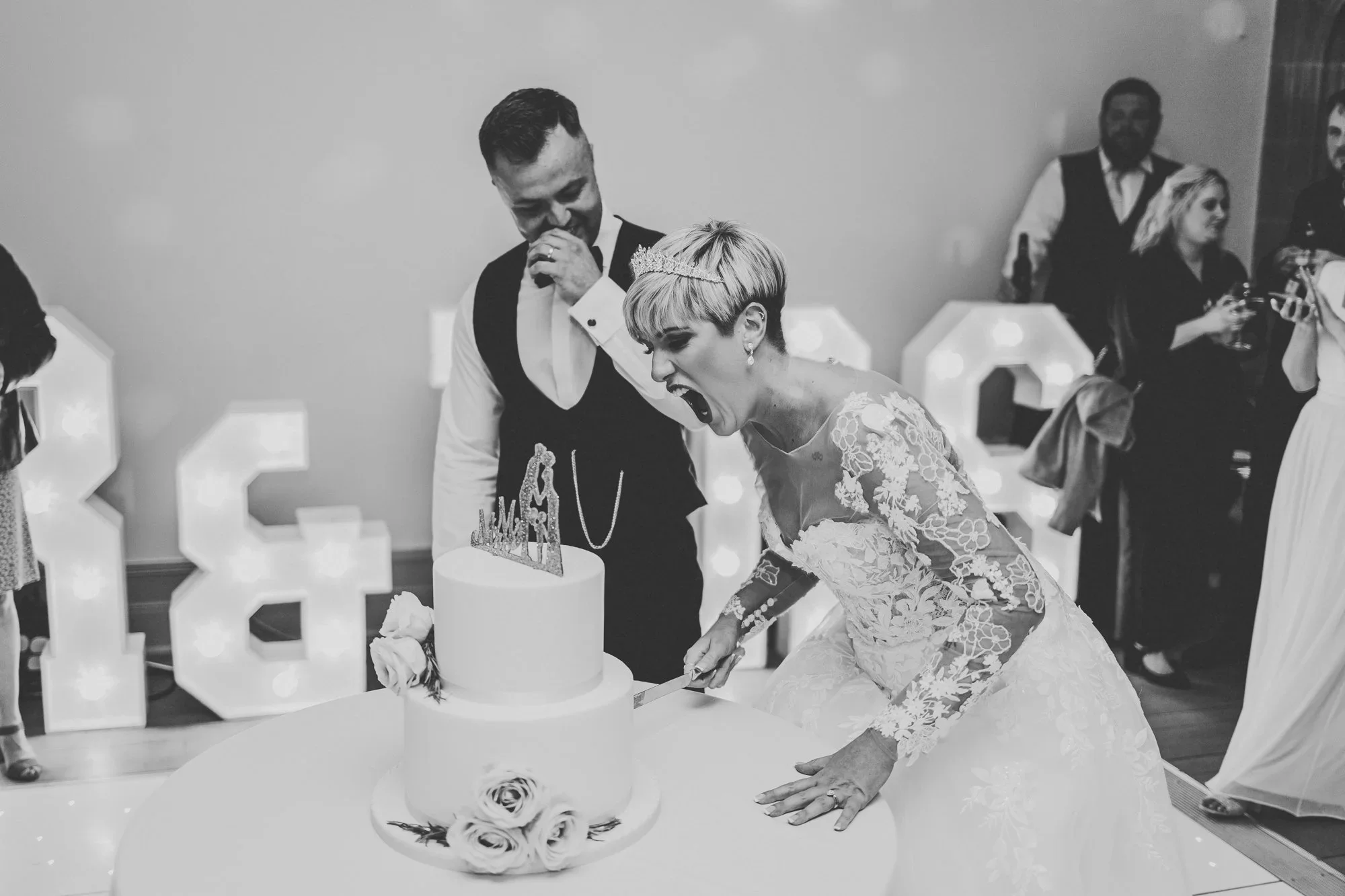A bride and groom cutting a wedding cake at their reception, with guests in the background. The bride is wearing a lace wedding dress and a tiara, and the groom is in a vest and white shirt.