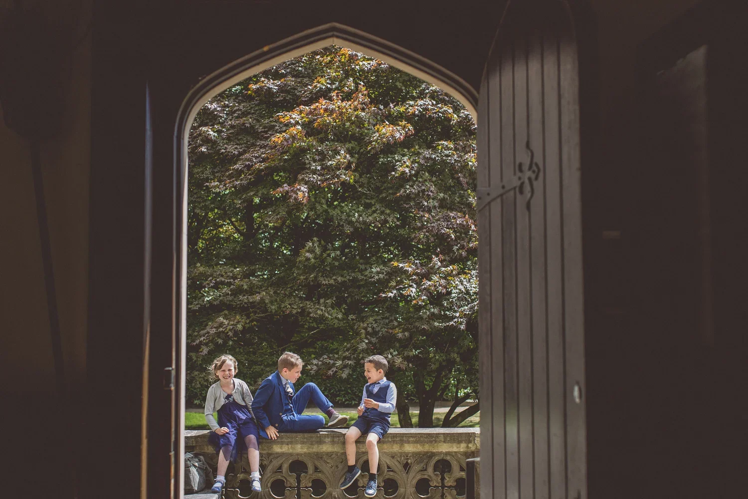 Three children sitting and talking on a stone bench outdoors, viewed through an arched doorway, with trees in the background.