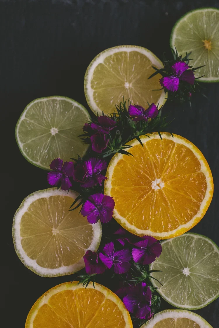 Slices of lemon, lime, and orange arranged on a dark surface with purple flowers.