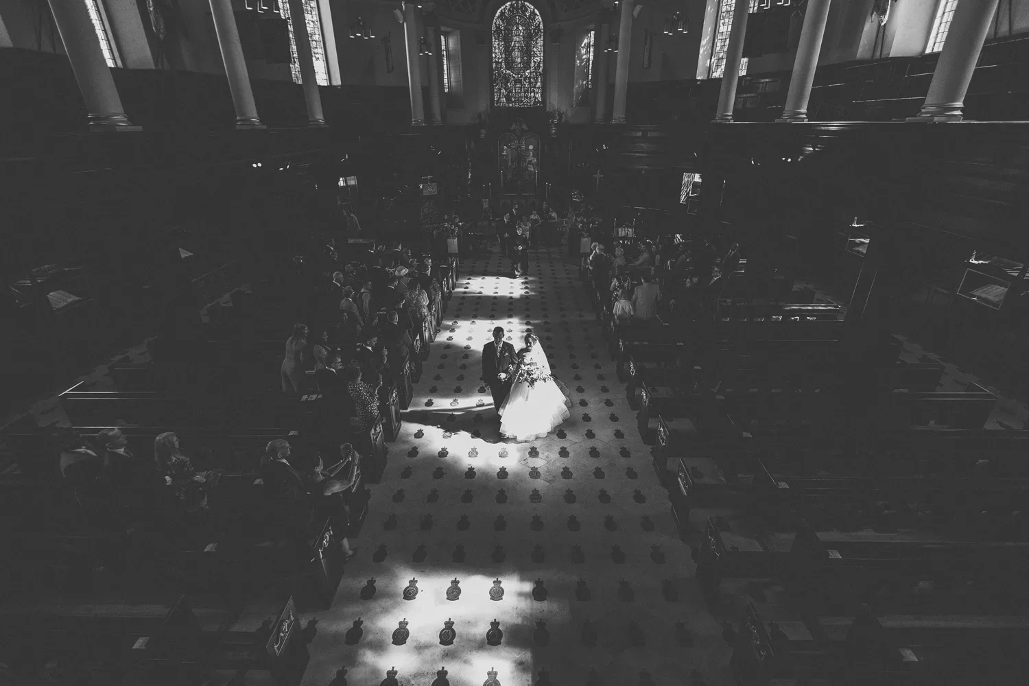 A black-and-white photo of a wedding ceremony inside a large church with high stained glass windows. The bride and groom are walking down the aisle, holding hands, surrounded by guests seated on wooden pews.
