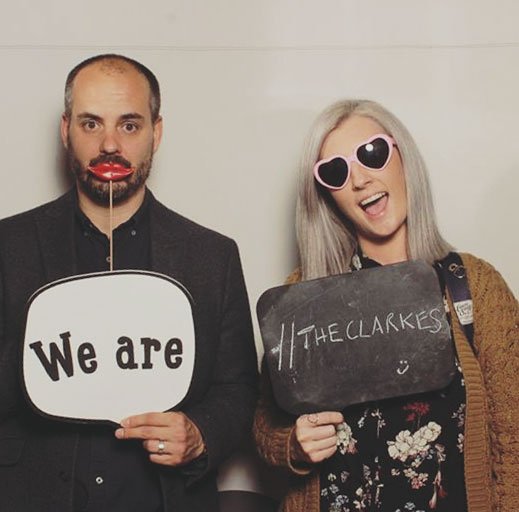 A man and woman standing against a plain wall. The man holds a speech bubble prop reading 'We are' and wears lipstick and a fake lip prop. The woman holds a blackboard with 'THECLARKES' and wears heart-shaped sunglasses, smiling.