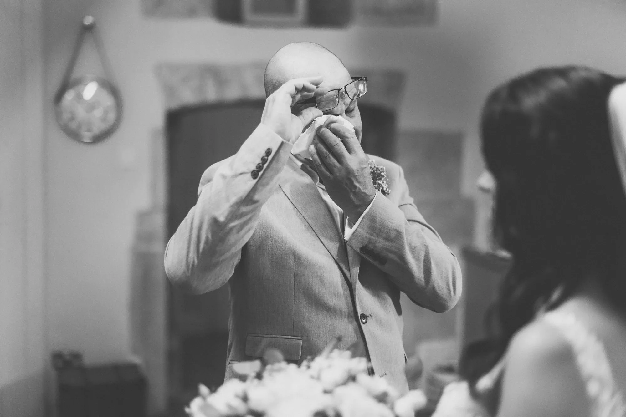 A bald man wearing glasses and a light-colored suit, holding a handkerchief to his face in a bathroom or dressing room, with blurred oval mirror on the wall behind him.