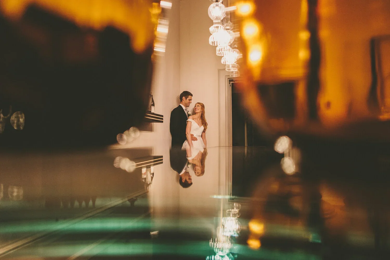 A couple, dressed in formal attire, standing near a wall with chandeliers hanging above, captured through a reflective surface with blurred edges.