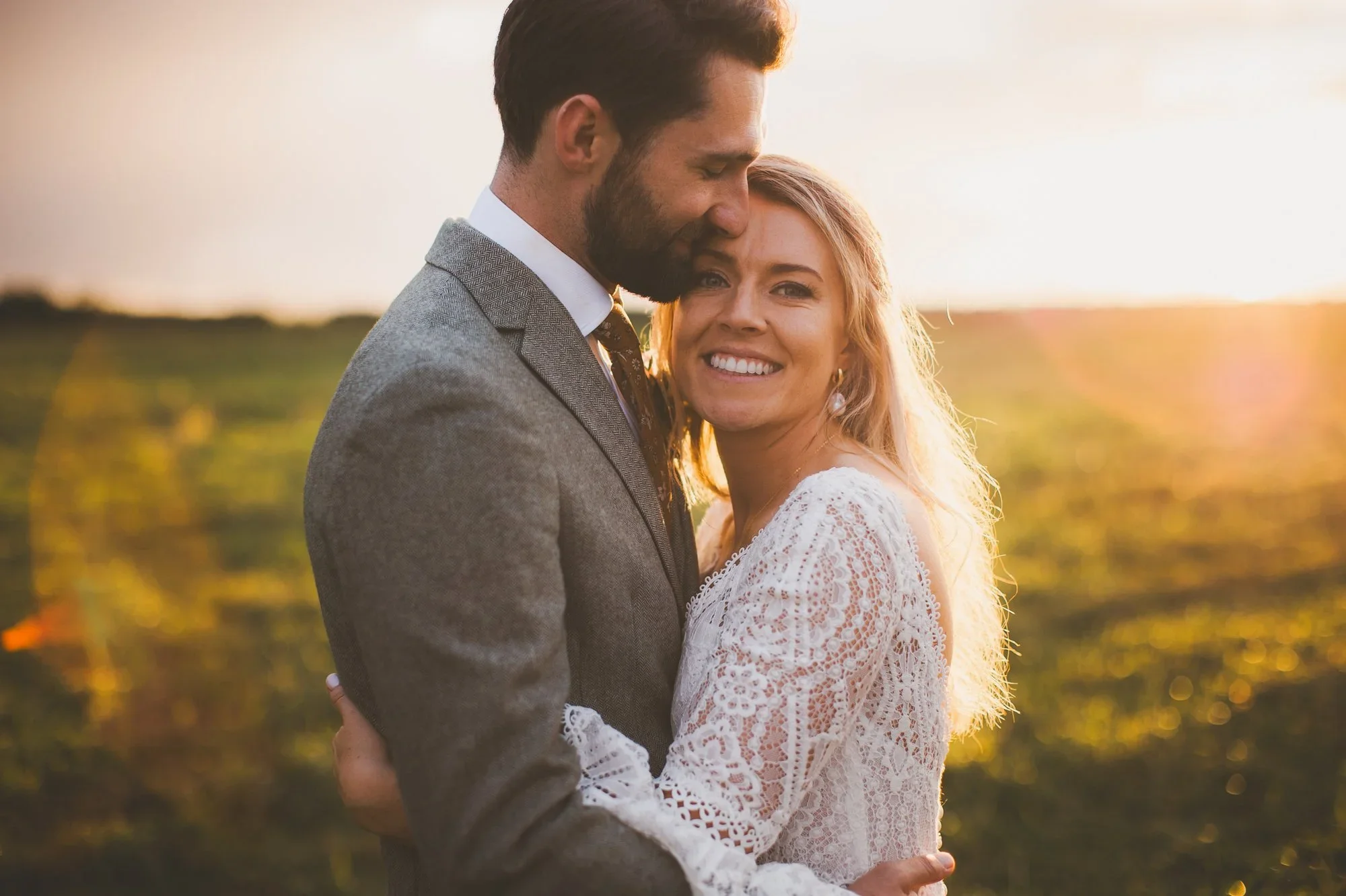 A smiling woman and a man in a gray suit embrace outdoors during sunset. The woman has blonde hair and wears a white lace dress, while the man has dark hair and a beard.