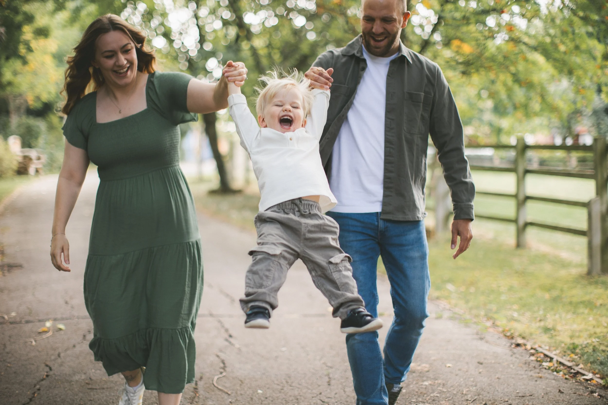 A young boy is being lifted in the air by two adults, his parents, as they walk on a park path surrounded by trees with leaves turning colors. The boy is smiling and laughing, wearing a white shirt and khaki pants. The woman on the left is wearing a 