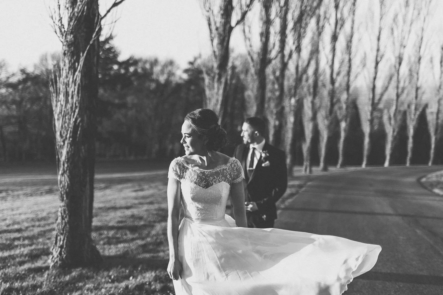 A black and white photo of a bride and groom outdoors, with the bride smiling and holding her dress as it flows in the wind, and the groom standing behind her, both dressed in wedding attire, near a winding road and leafless trees.