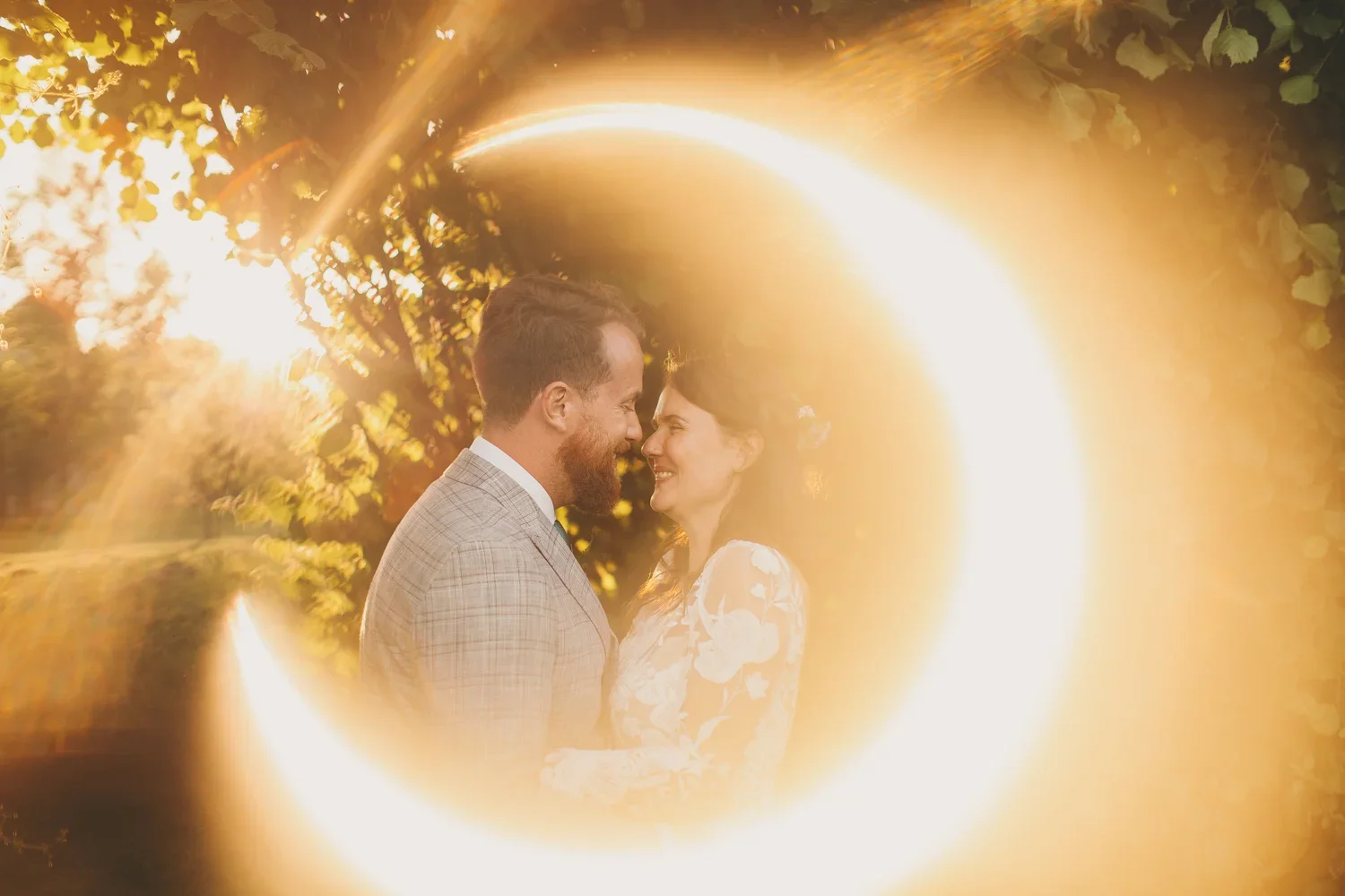 A couple standing close together outdoors during sunset, with their foreheads touching and smiling at each other, surrounded by warm light and a golden glow.