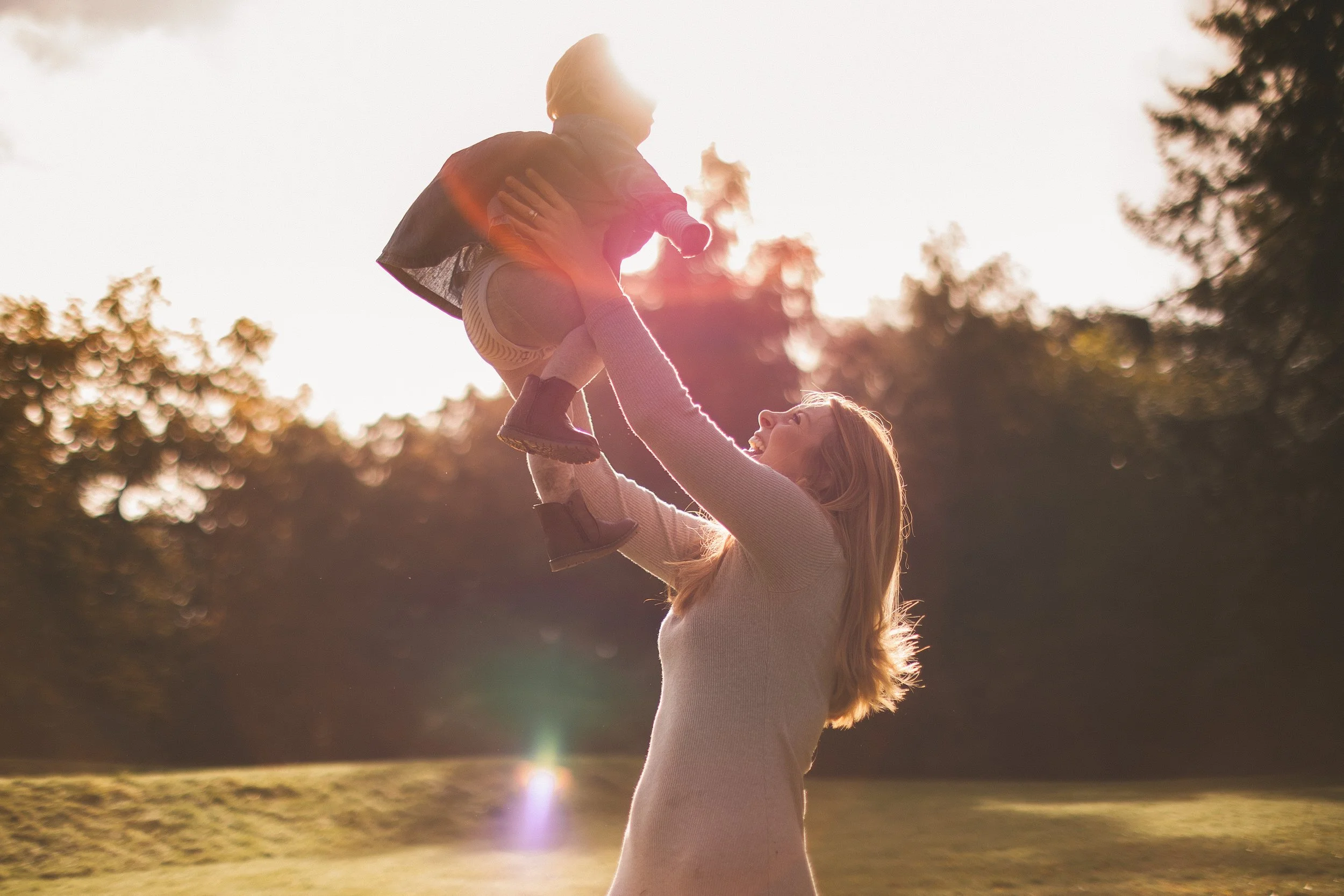 A woman lifting a child in the air outdoors during sunset, with trees in the background.