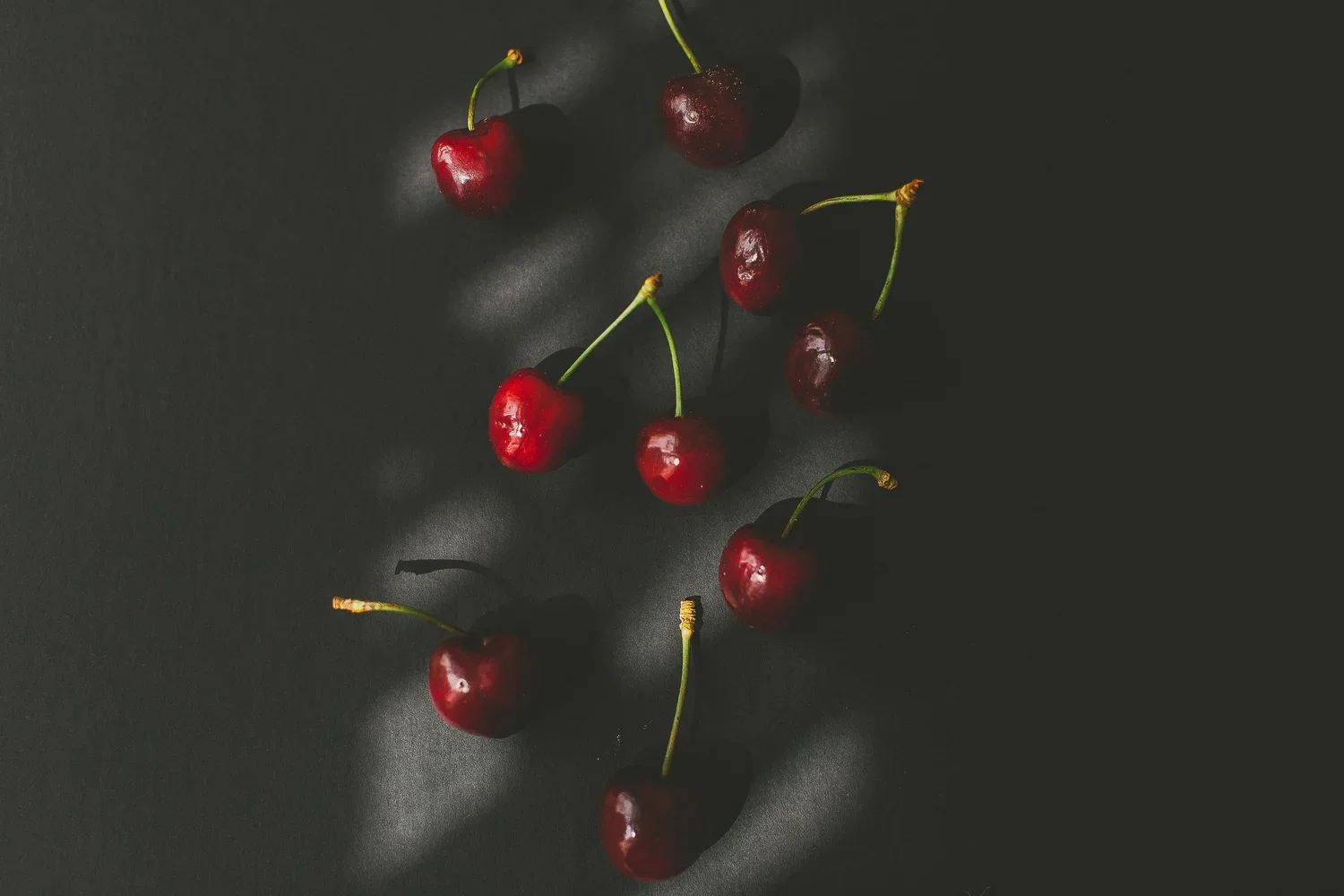 A handful of cherries with green stems scattered on a dark surface, casting shadows.